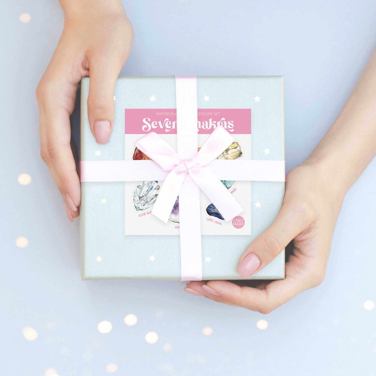 a woman's hands holding a blue gift box with a white ribbon with the seven chakras watercolor crystal sticker sheet