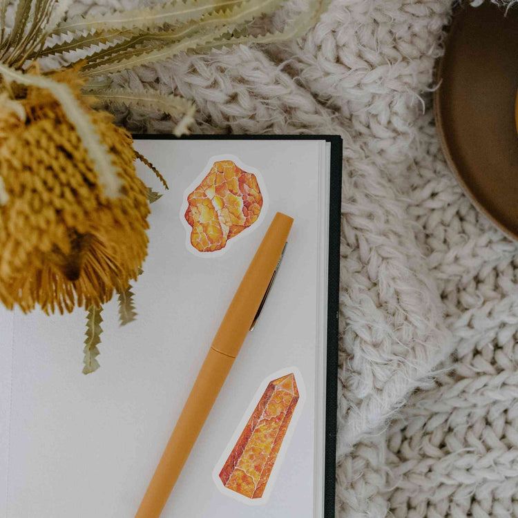 a notebook with orange calcite stickers on it from sacral chakra watercolor crystal sticker set next to a dried plant