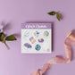 a watercolor crystal sticker sheet for the crown chakra on a purple surface next to a flower