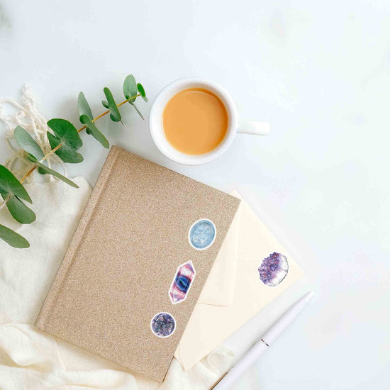 a cup of coffee and a notebook with fluorite, amethyst, calcite, and polite stickers on a table